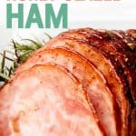 Ham sits on a white dish. Juices from the glaze surrounds the ham. A text overlay reads "Slow Cooker Honey Glazed Ham."