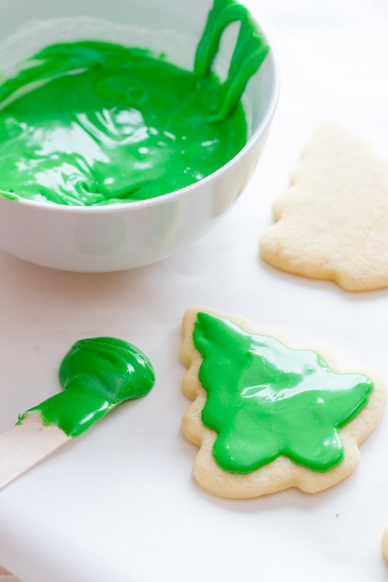Cut-out cookies shaped like evergreen trees sit next to a bowl of green frosting. One cookie has some green icing on it already.