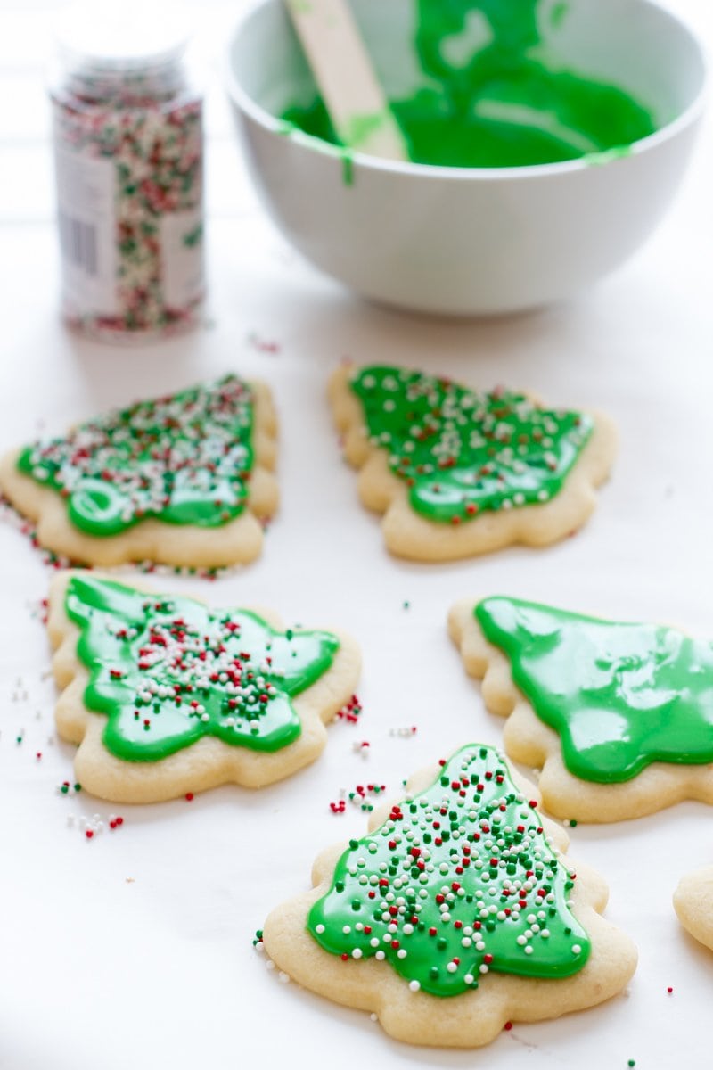 Decorated Christmas tree sugar cookies with green frosting rest on a white background with a bowl of sugar cookie icing in the back.