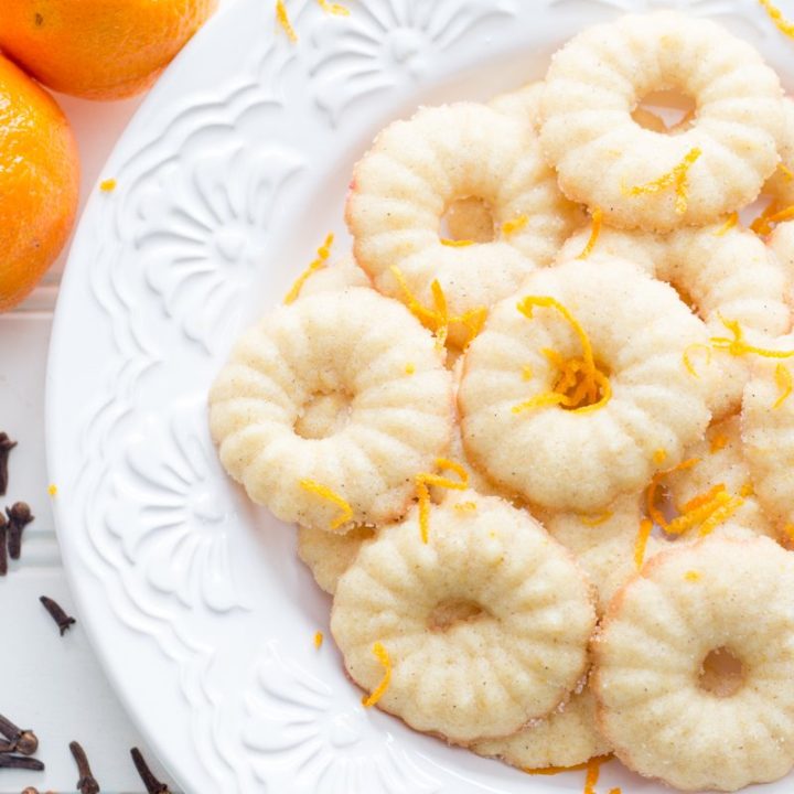 Clementine and Clove Spritz Cookies on a white plate with clementines and cloves nearby