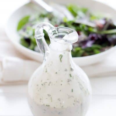 Glass bottle filled with healthy ranch dressing sits in front of a white bowl filled with salad.