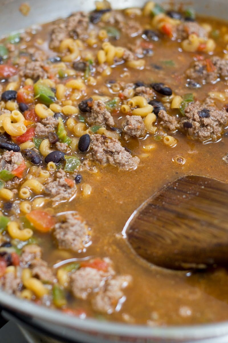 Pasta, black beans, beef and other ingredients cook together in a silver pan. A wooden spoon stirs the mixture.
