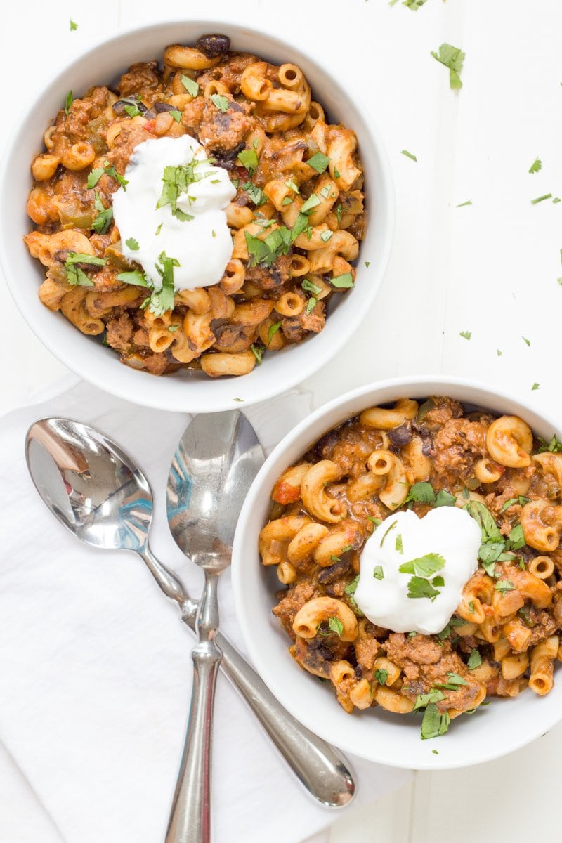 Two bowls of the taco macaroni skillet sit on a white background. Two spoon sit nearby on a white napkin.