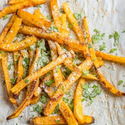 A pile of baked pumpkin fries dusted with chopped herbs and parmesan cheese sits on a parchment paper lined baking sheet.