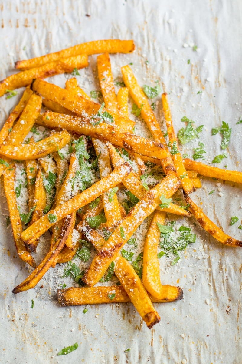 A pile of baked pumpkin fries dusted with chopped herbs and parmesan cheese sits on a parchment paper lined baking sheet.