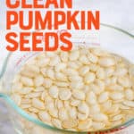 Raw pumpkin seeds soak in a large glass measuring cup full of water. The measuring cup is on a white marble countertop. A text overlay reads "The Easy Way to Clean Pumpkin Seeds."