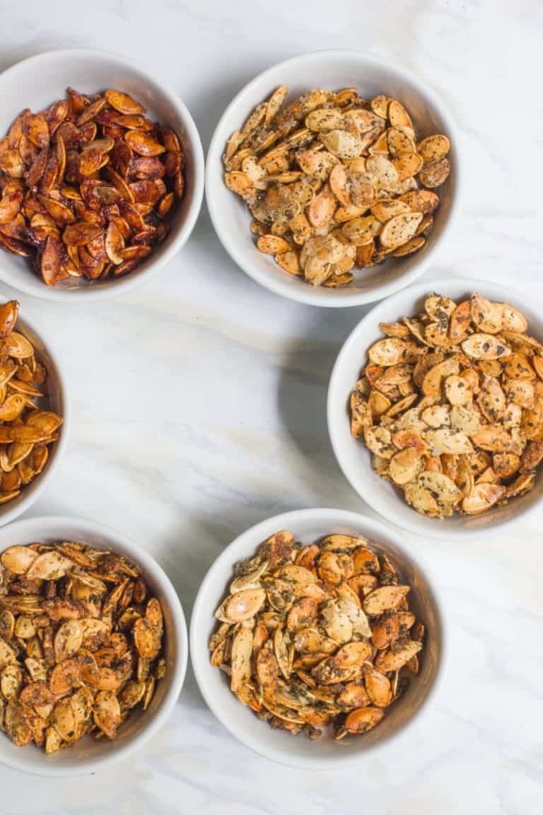 Six small white bowls filled with different flavors of roasted pumpkin seeds. Bowls are placed on a white marble countertop.