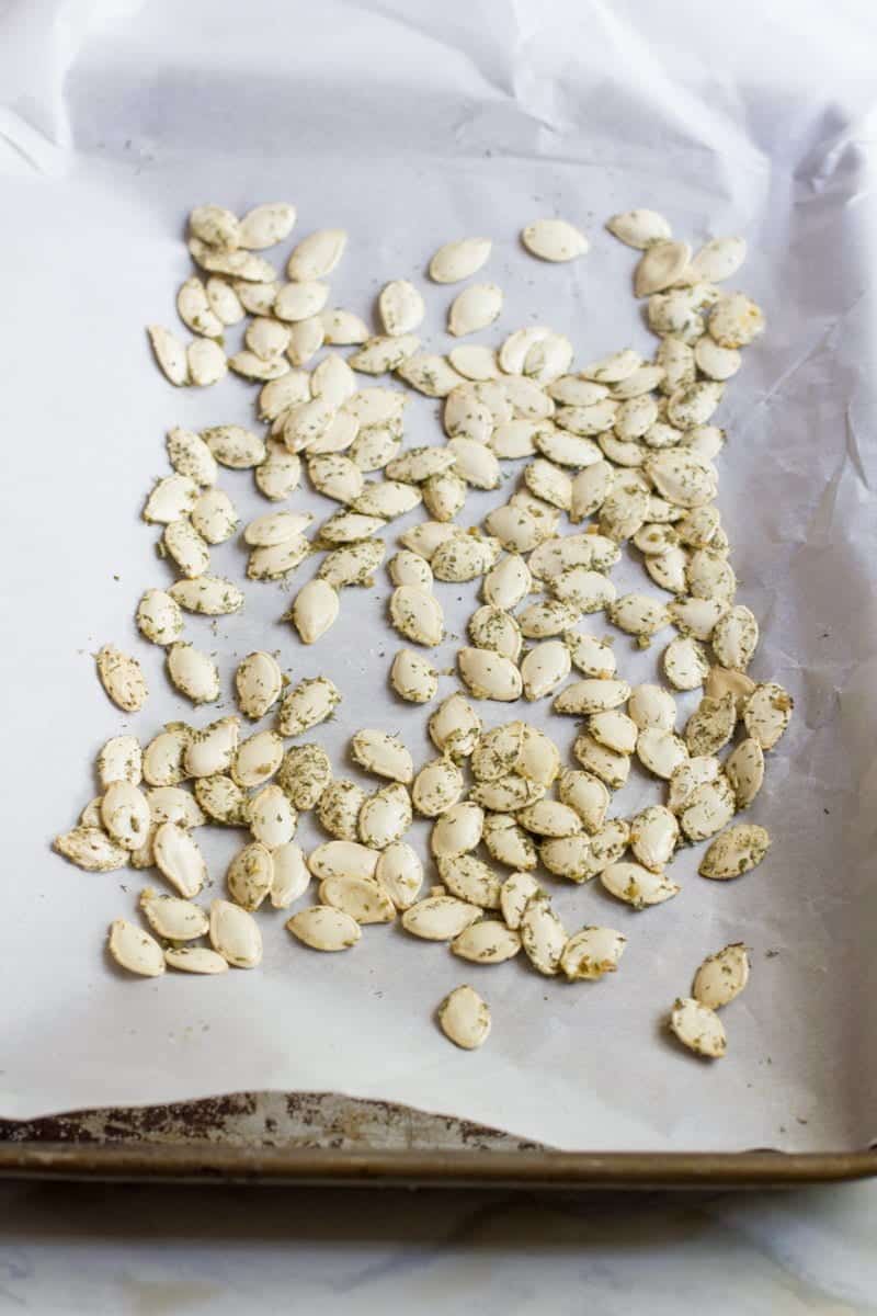 Unroasted, seasoned pumpkin seeds are spread out on a parchment paper lined baking sheet.
