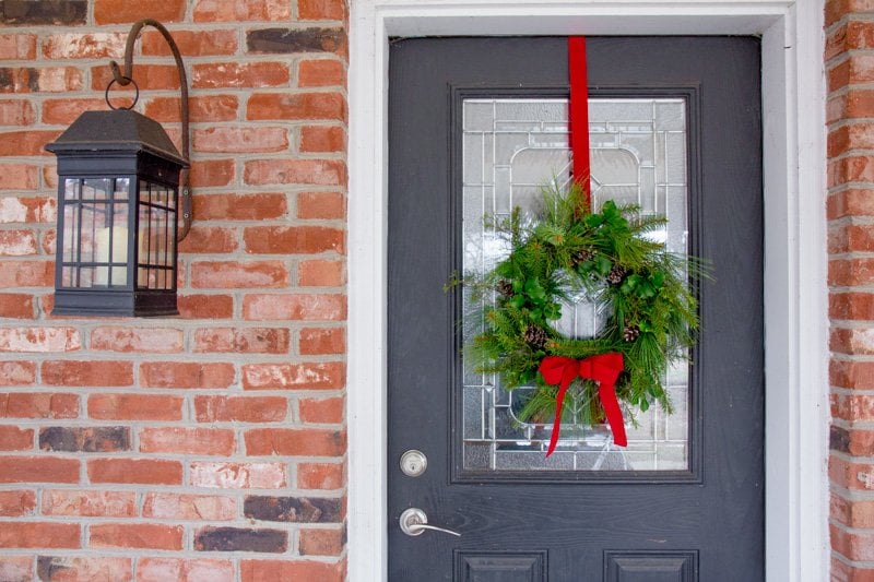 Evergreen wreath hanging from a red ribbon on a front door of a brick house.