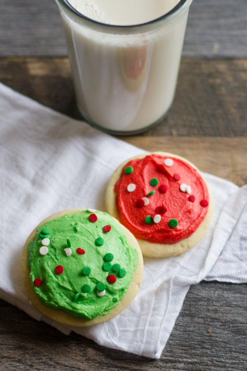 2 Lofthouse Sugar Cookies - one with green frosting and one with red - on a white napkin with a glass of milk