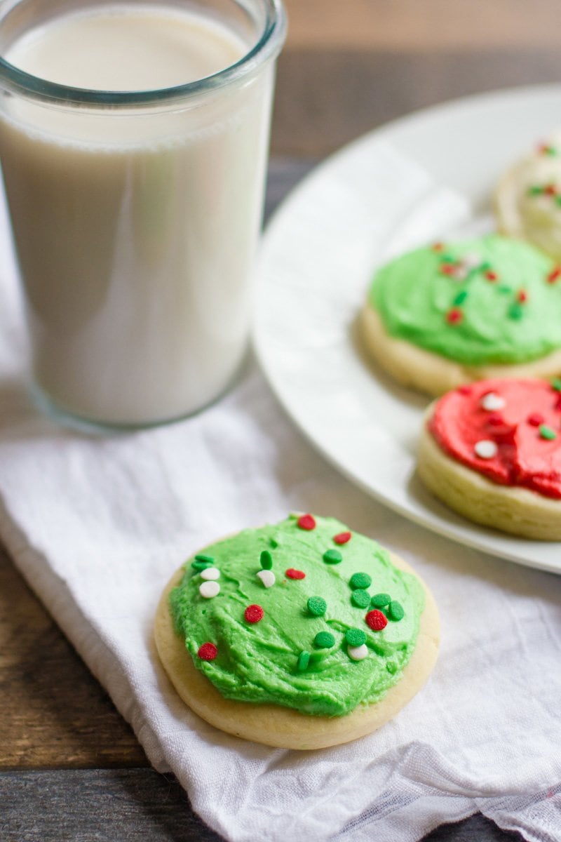Lofthouse Sugar Cookie with green frosting and sprinkles, next to a glass of milk and a plate of cookies