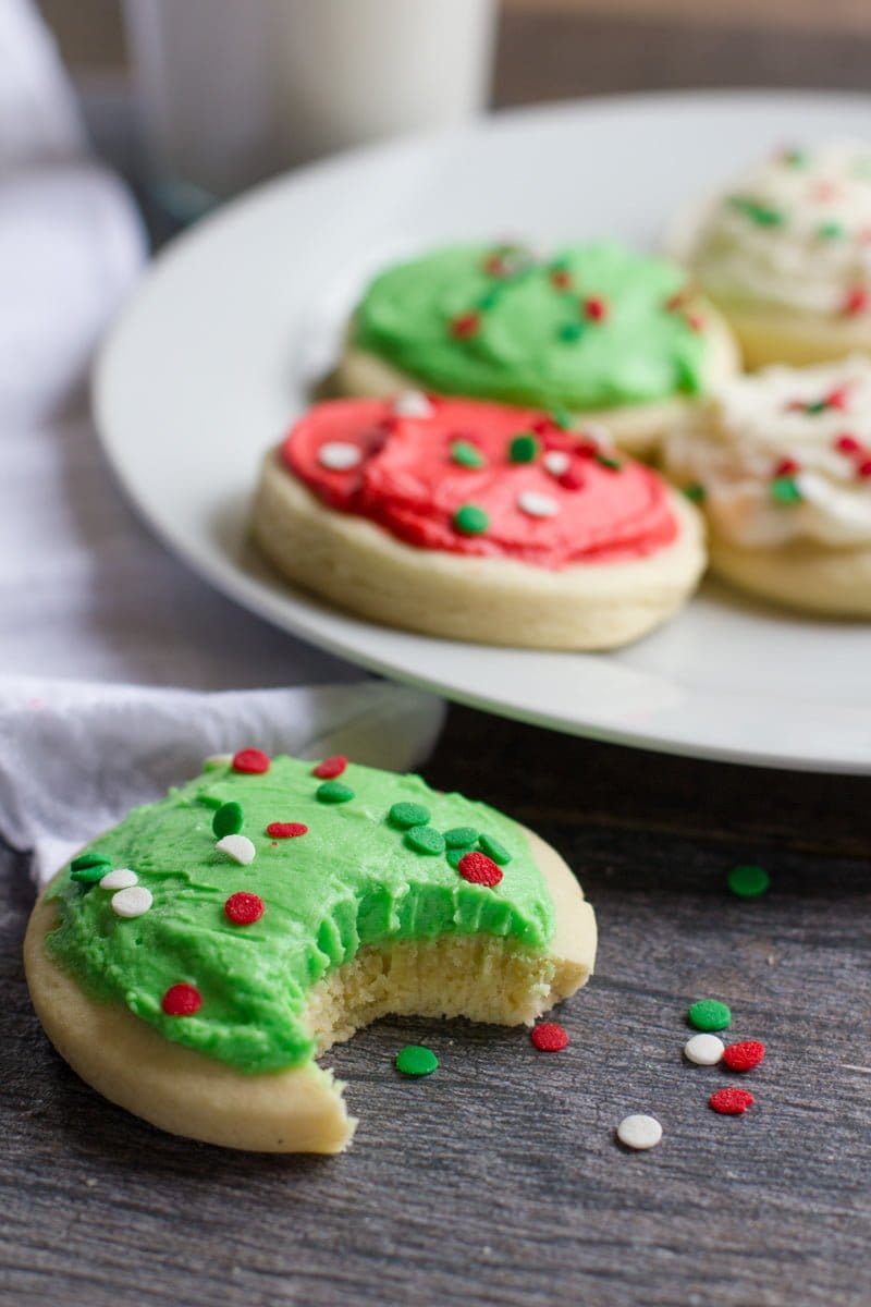Lofthouse Sugar Cookie with green frosting and sprinkles with a bite taken out of it, in front of a plate of other cookies