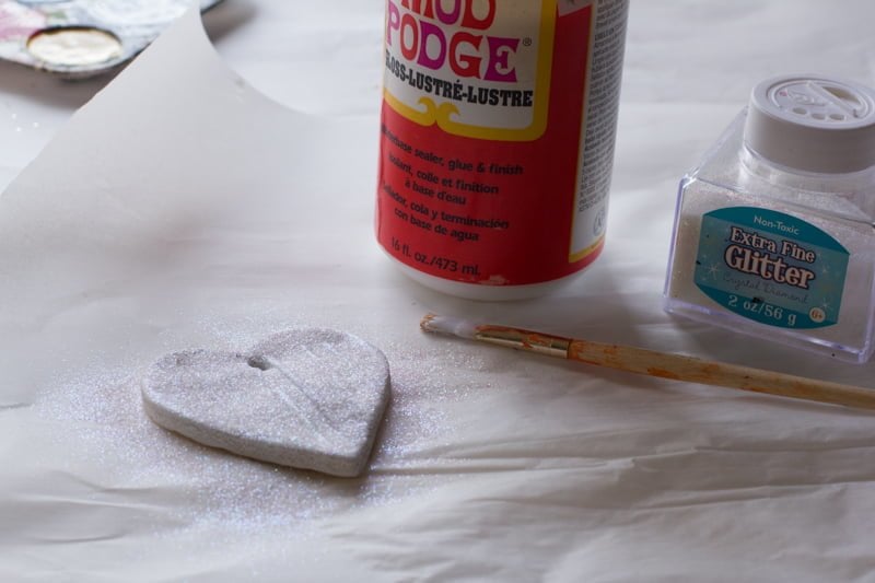 A salt dough ornament covered in white glitter sits on a white background with a bottle of Mod Podge and glitter in the background.