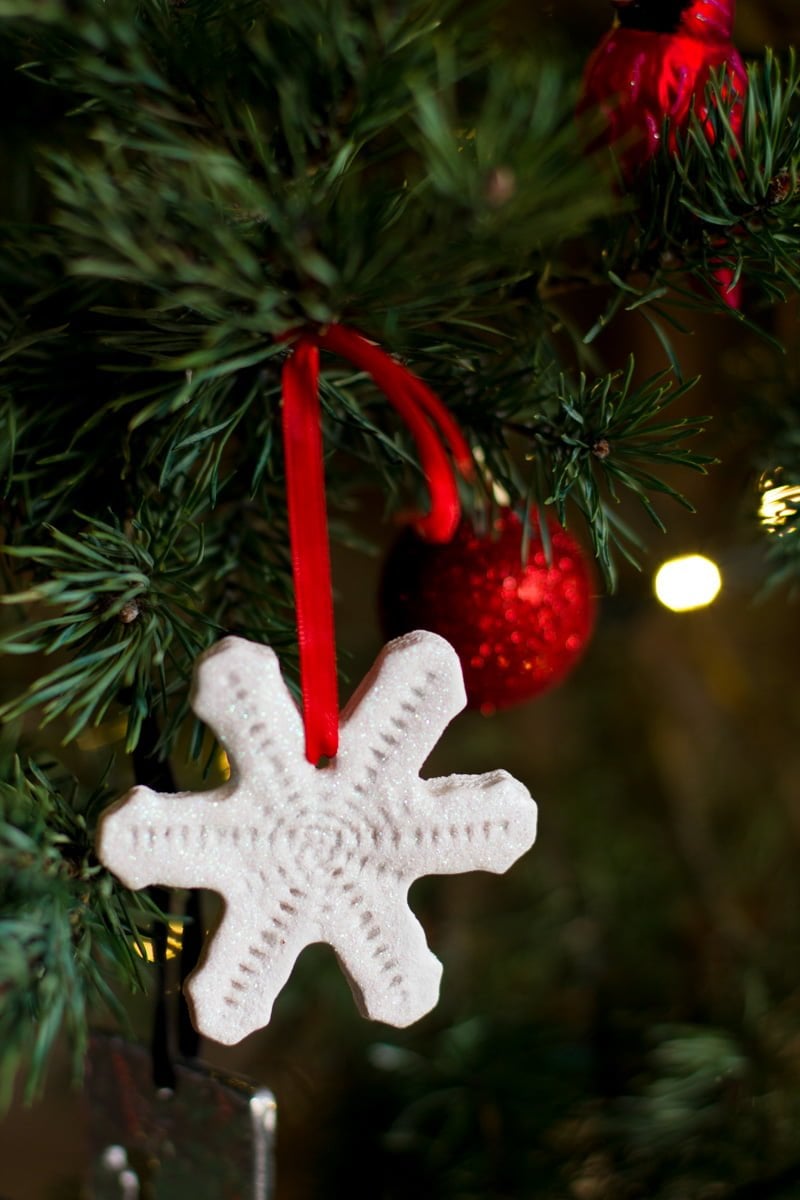 A sparkly snowflake shaped salt dough ornament hangs from a Christmas tree with a red ribbon. Christmas lights and red sparkly ornaments are blurry in the background.