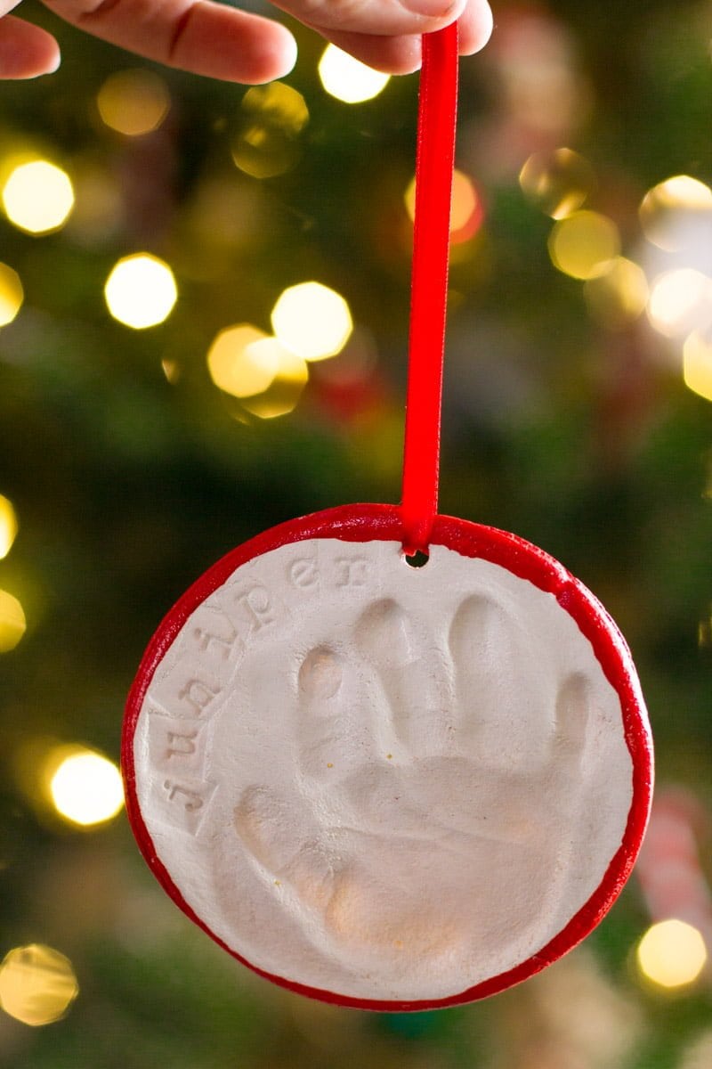 A hand holds a salt dough handprint ornament hanging from a red ribbon. The background is a blurry Christmas tree with lights.