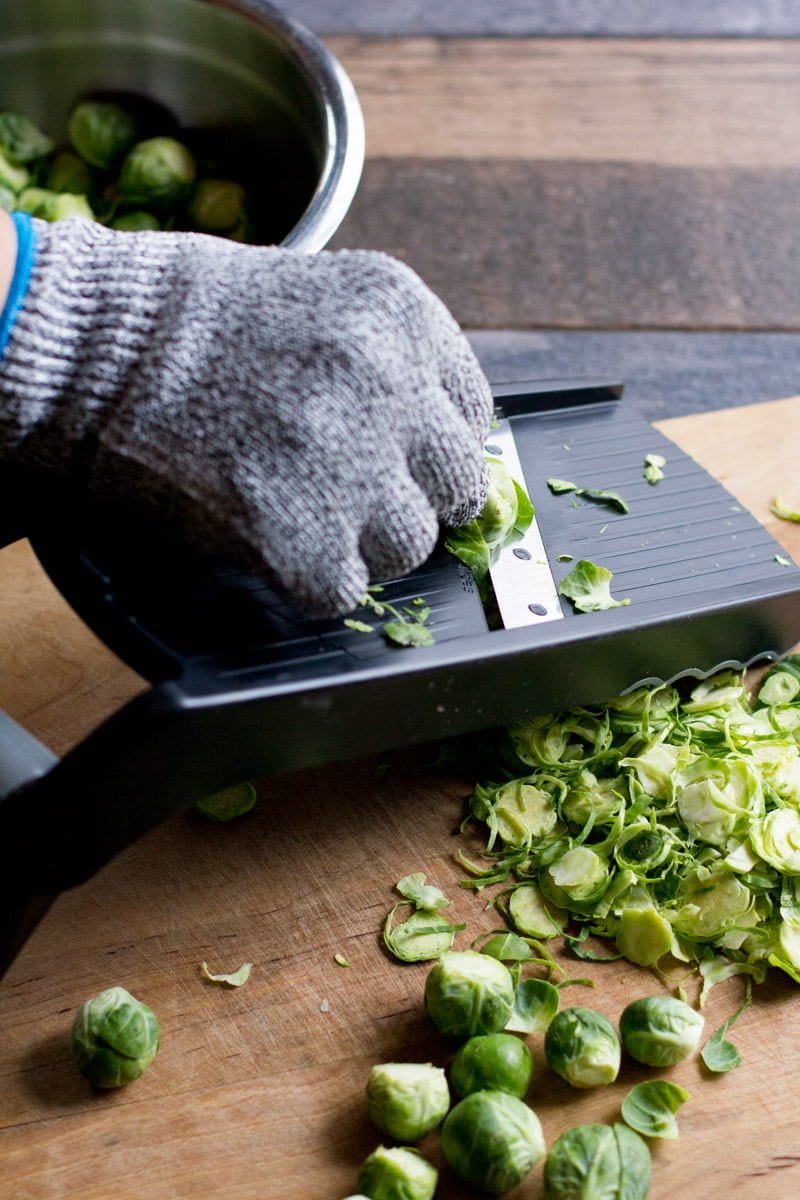 A gloved hand uses a mandoline to slice Brussels sprouts.