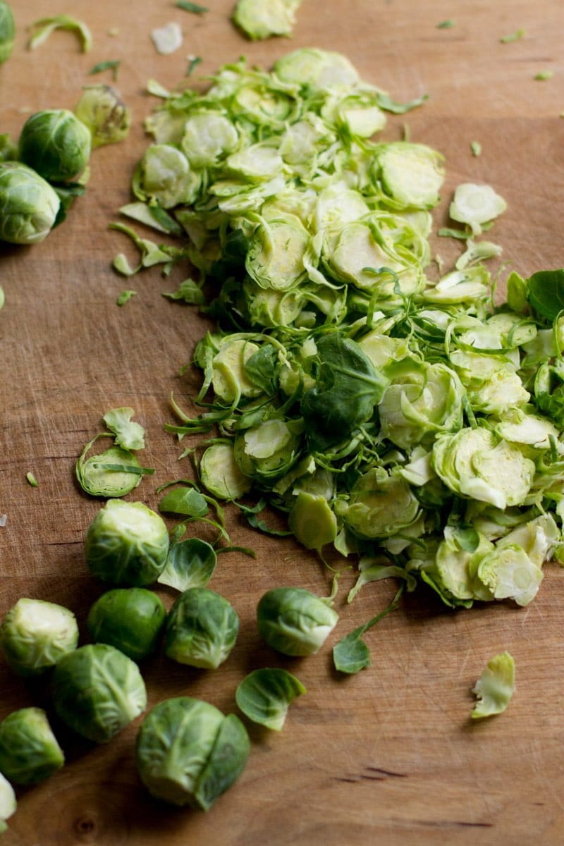 Brussels sprouts are piled on a wooden cutting board. Some of the sprouts are already shaved, and others are whole.