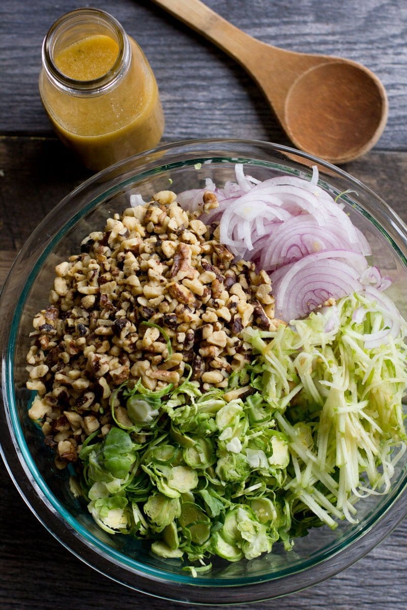 Shredded Brussels sprouts, chopped walnuts, sliced onions, and sliced apples in a glass bowl, ready to be mixed together. A wooden spoon and a jar of dressing sit nearby.