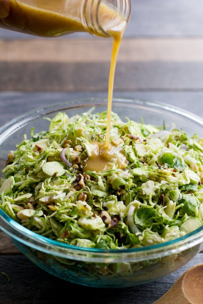 Salad dressing pours from a glass jar and into a glass bowl of shredded Brussels sprouts and other ingredients
