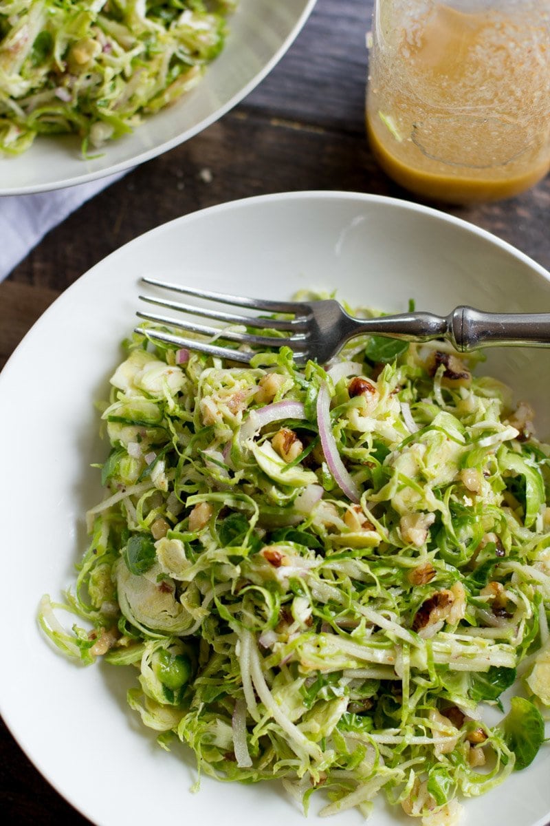 A white plate filled with Shaved Brussels Sprout Salad with Apples and Walnuts sits next to an almost-empty bottle of homemade dressing.