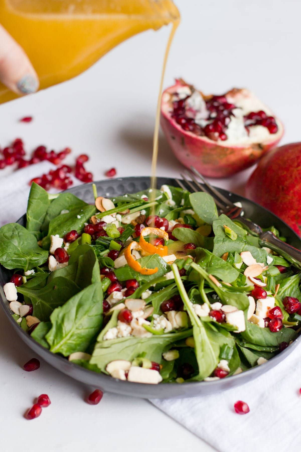 Pouring clementine vinaigrette over a baby spinach salad topped with sliced almonds and pomegranate arils.
