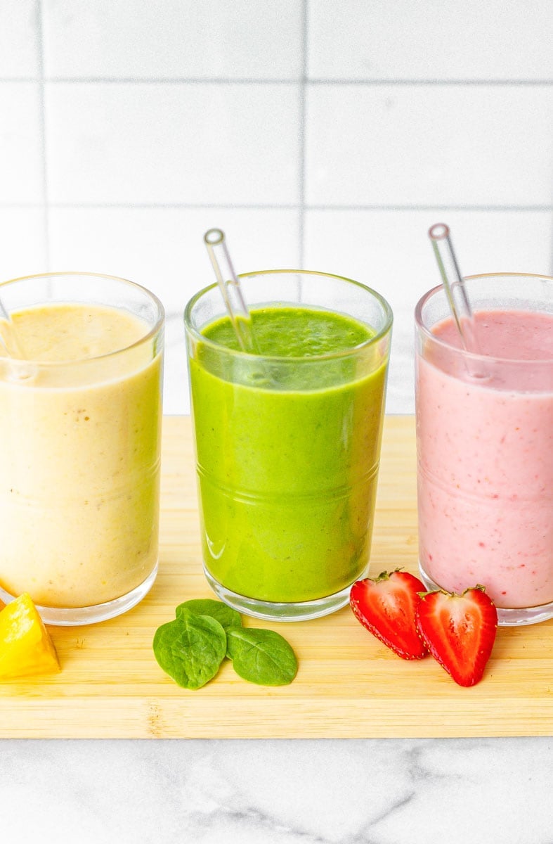 Three fruit smoothies are lined up in clear glasses with glass straws.