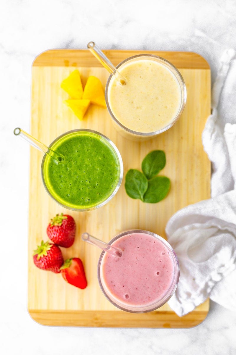 Three fruit smoothies sit on a cutting board, with fresh fruit and spinach around them.