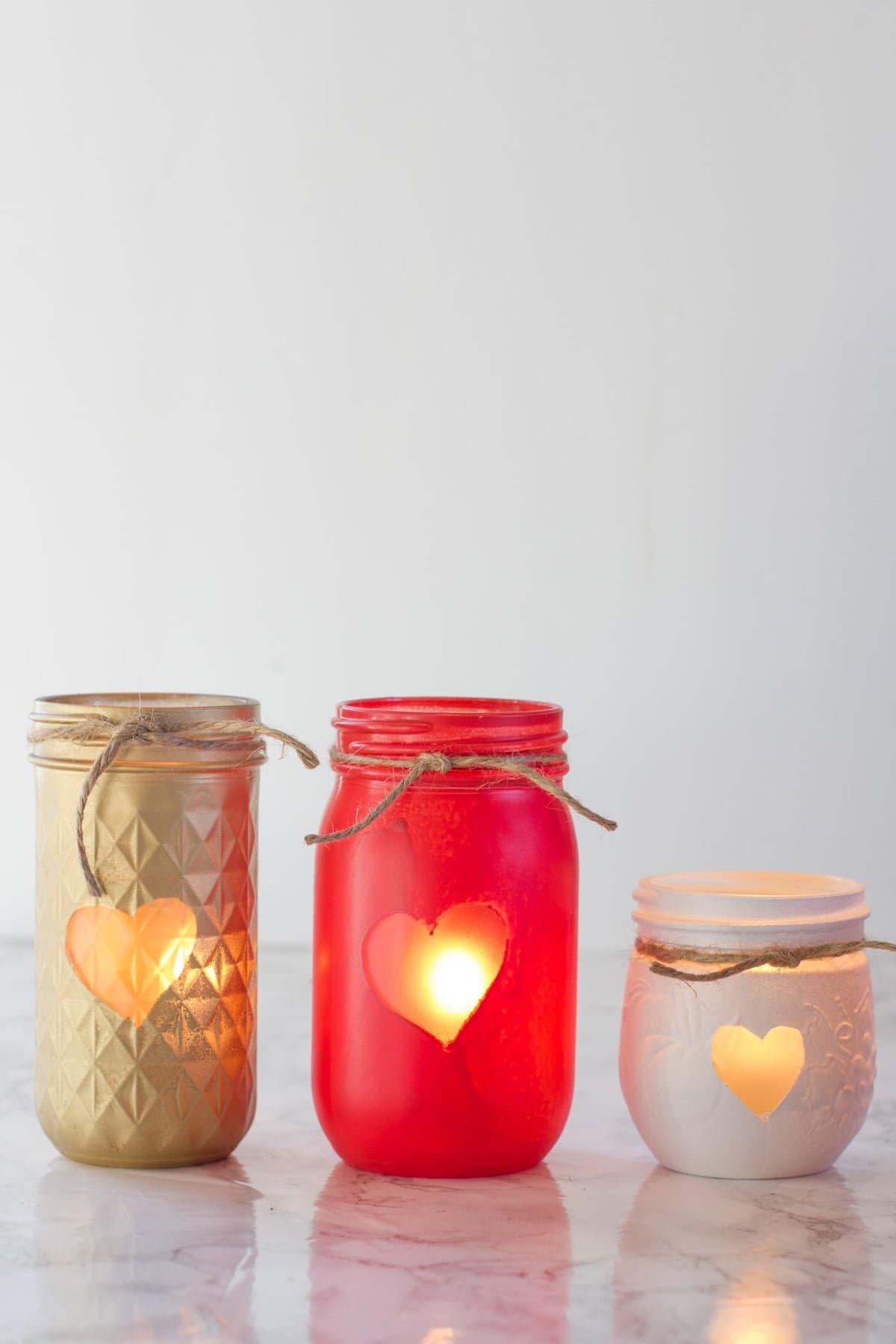 Three Painted Mason Jar Votive Holders, each with a heart and a piece of twine. The jars are gold, red, and white.