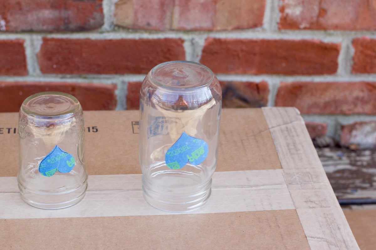 Two mason jars with painter's tape hearts sitting upside down on a piece of cardboard