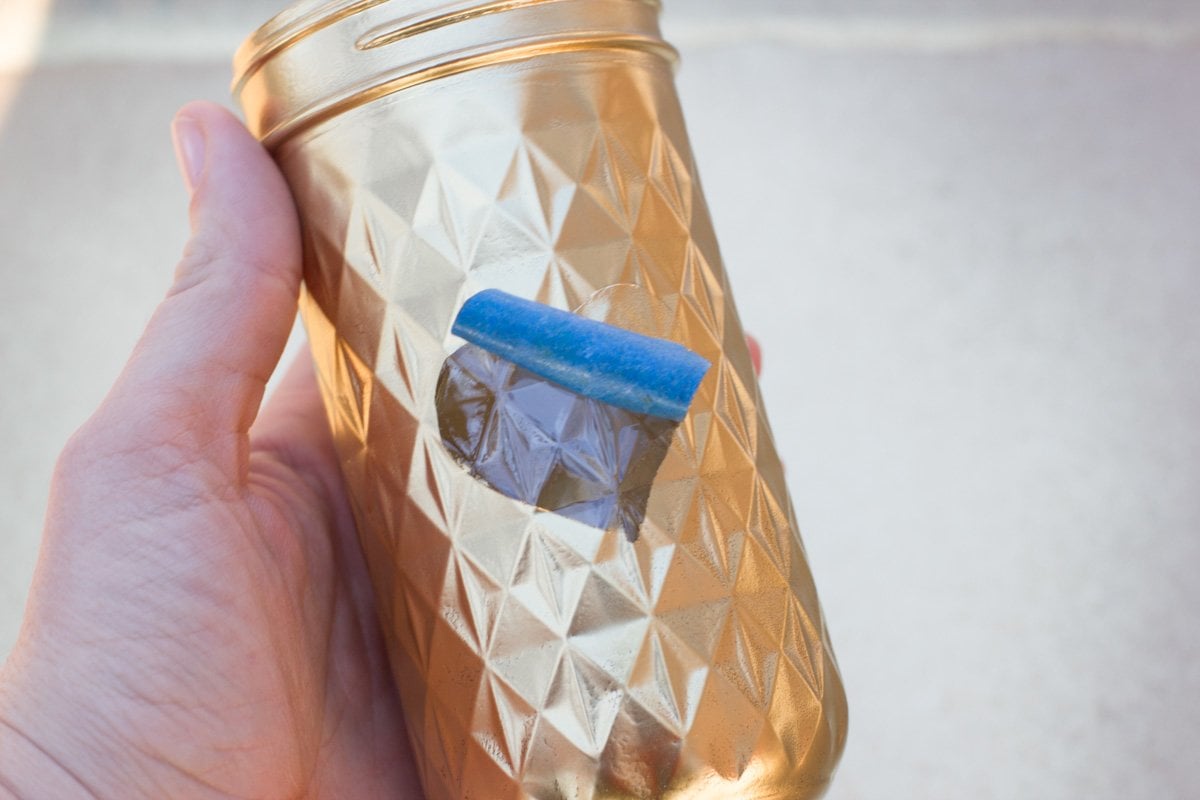 Hand holding a mason jar that has been spray painted gold around a heart shape, with a painter's tape heart partially peeled off