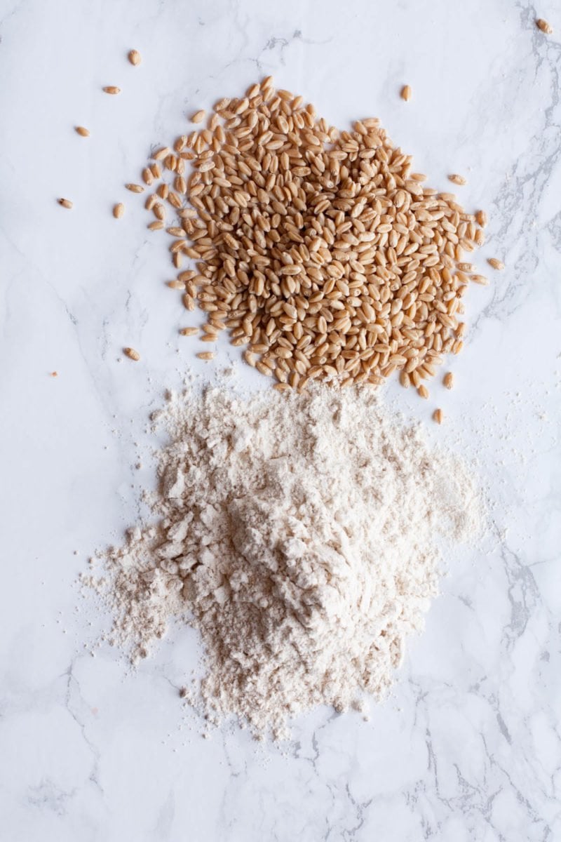 Wheat kernels and ground flour sit in two piles on a marble background.