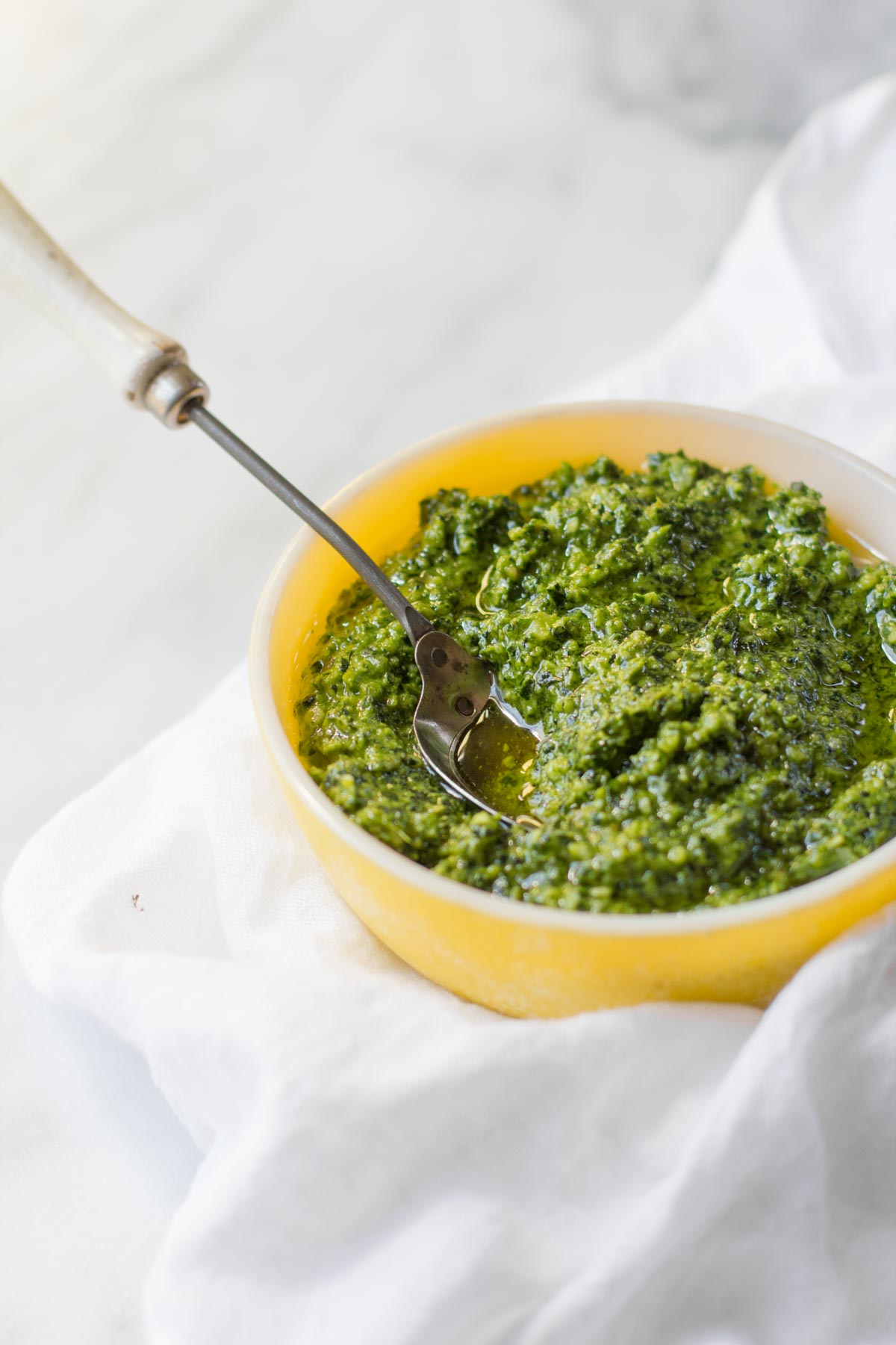 A bowl of Kale and Walnut Pesto sits with a spoon, on a white background.
