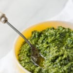 A bowl of kale and walnut pesto sits on a white background. A text overlay reads "Kale + Walnut Pesto"