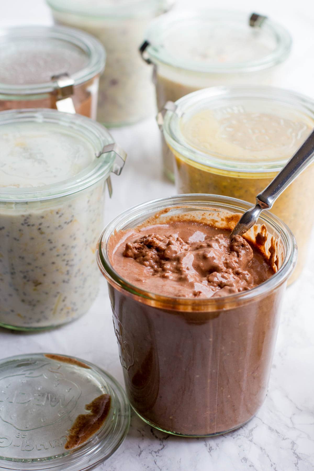6 glass jars full of different flavors of overnight oats. All have lids, except for one chocolate flavor, which is open and has a spoon sticking out of it.