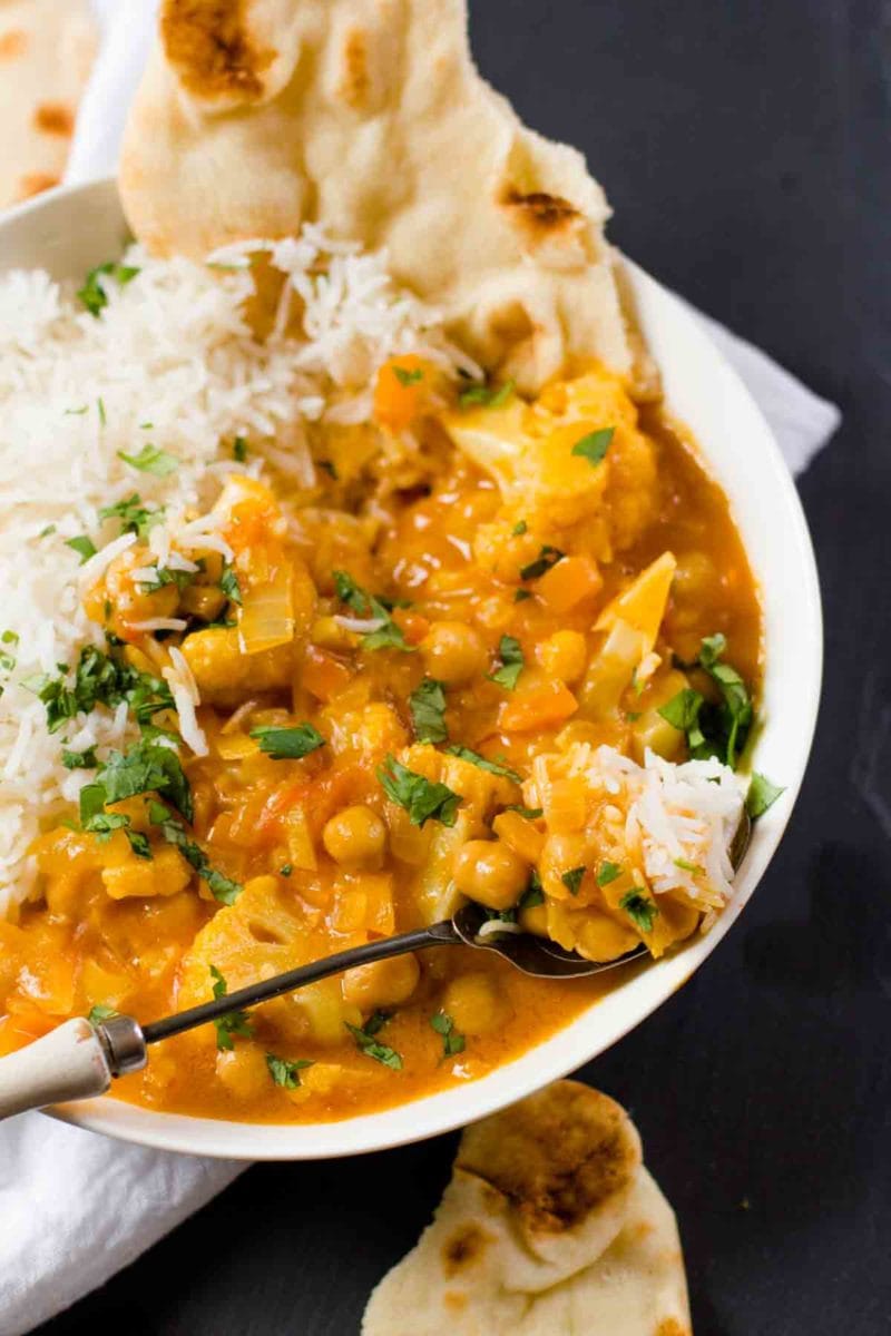 Spoon full of Cauliflower and Chickpea Curry resting on a bowl of curry, rice, and naan