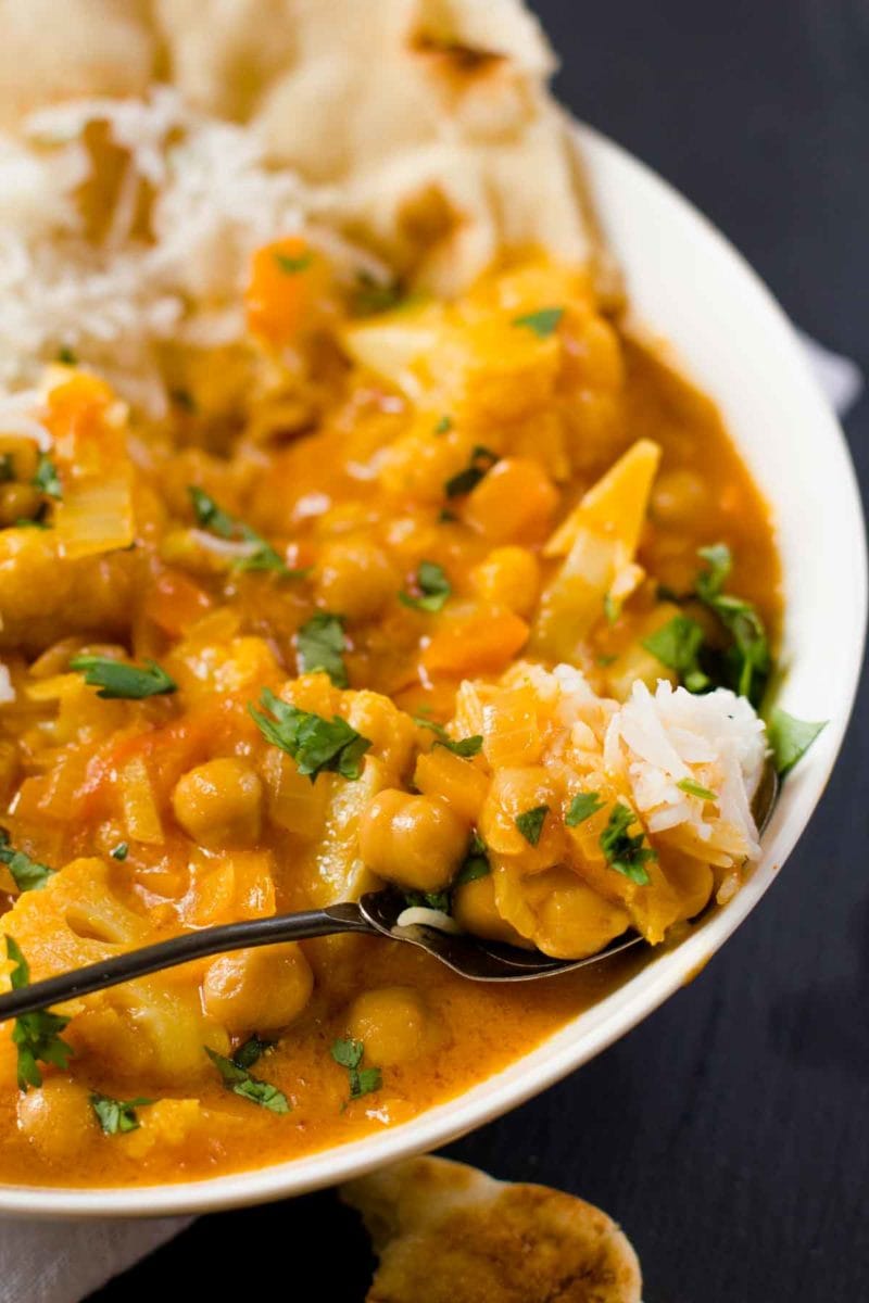 Spoon full of white rice and chickpea and cauliflower curry on a spoon that is resting on the edge of a white bowl of curry.