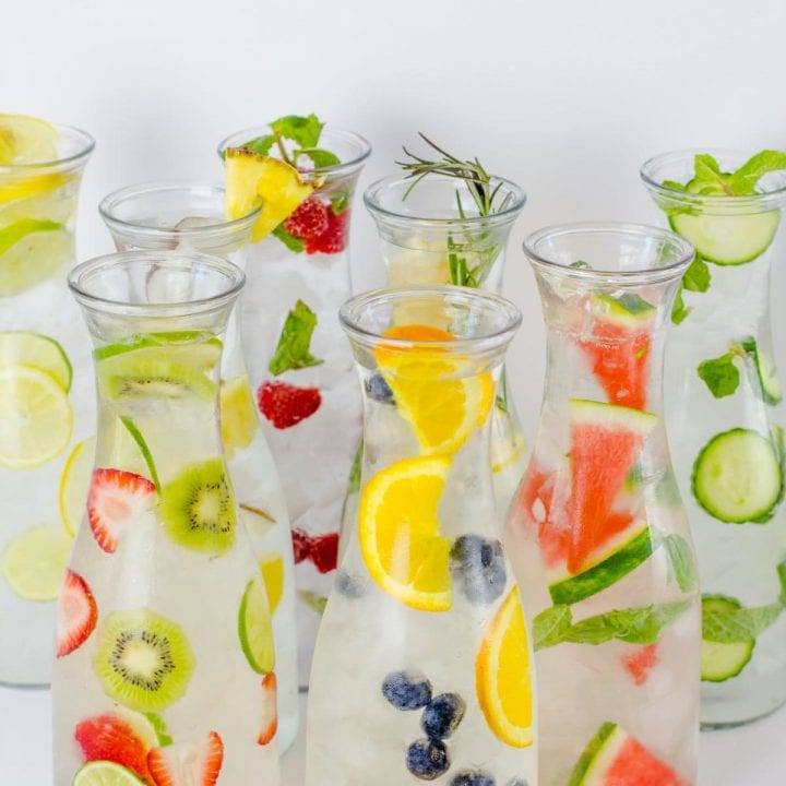 Eight glass carafes filled with different flavors of fruit-infused water are clustered together.
