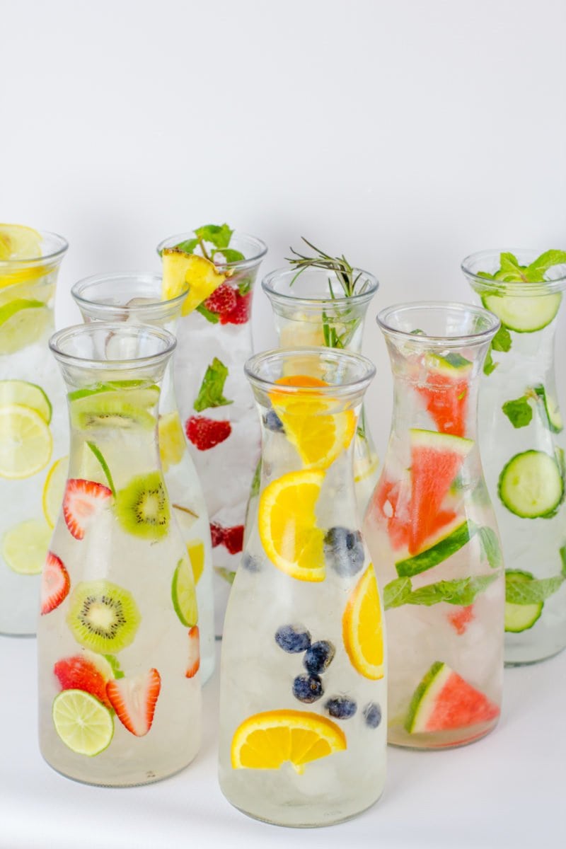 Eight glass carafes filled with different flavors of fruit-infused water are clustered together.