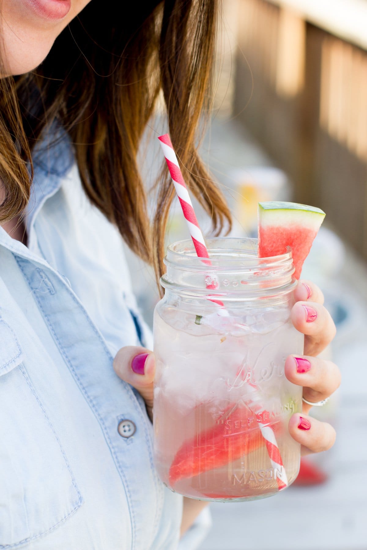 Close-up of a woman holding a mason jar filled with watermelon infused water and a striped paper straw.