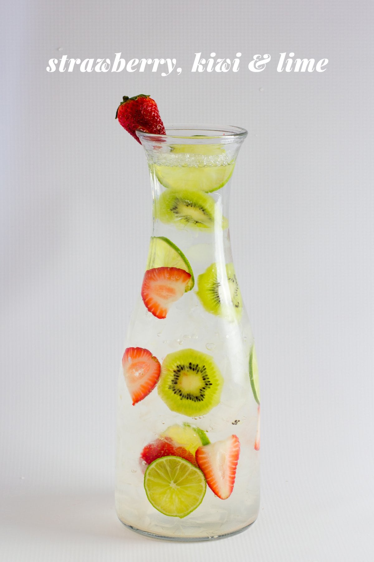 Strawberry, kiwi, and lime infused water is displayed in a glass carafe. A text overlay reads, "Strawberry, Kiwi, & Lime."