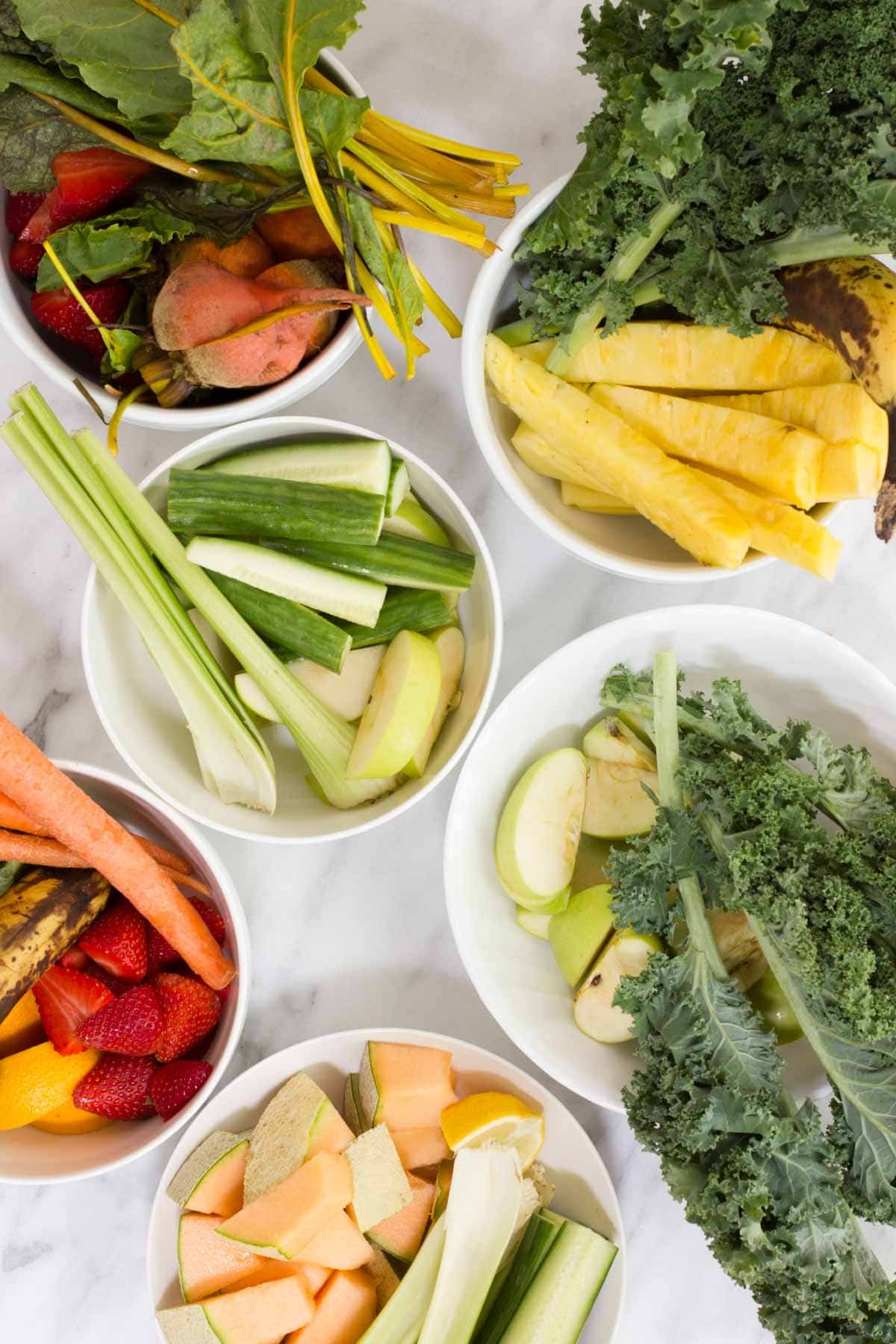 Five white bowls filled with various fresh fruits and vegetables for juice recipes, including kale, pineapple, carrots, strawberries, cucumbers, apples, celery, and melon.