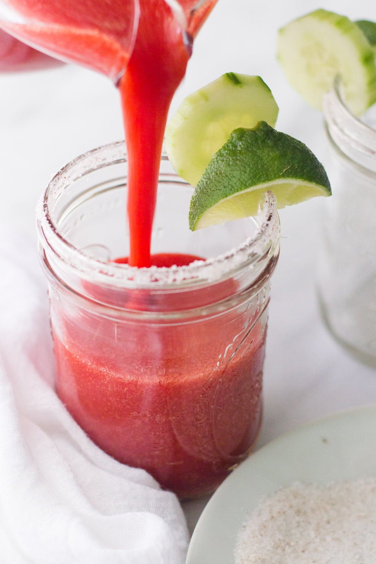 Strawberry margarita pours into a salt-rimmed jar garnished with a cucumber slice and lime wedge.