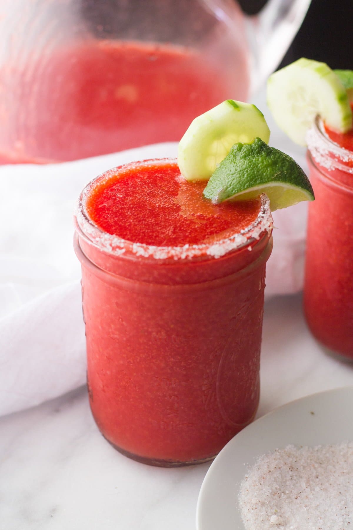 A frozen strawberry margaritas with cucumber stands on a counter with a second drink beside it and a pitcher behind it.