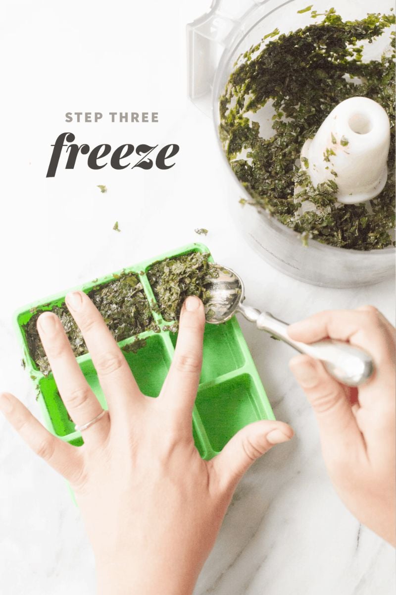 Hands spoon fresh basil pureed with olive oil out of a food processor and into an herb freezing tray. A text overlay reads "Step Three: Freeze."