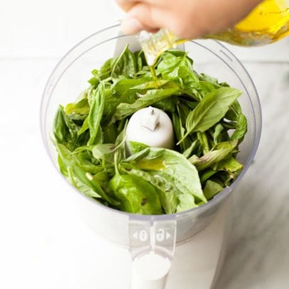 Hand pours olive oil into a food processor filled with fresh basil leaves.