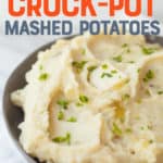Mashed potatoes in a grey bowl, garnished with a pat of butter and chopped chives. A text overlay reads "The Best Ever Crock-Pot Mashed Potatoes."