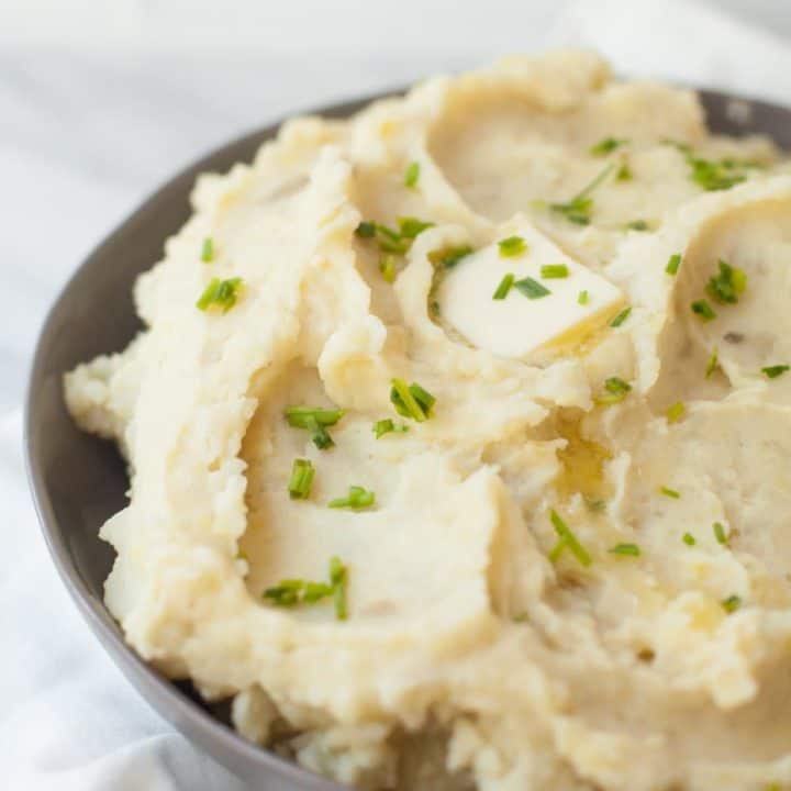 Tight view of bowl of fluffy slow cooker mashed potatoes on white countertop. Mashed potatoes are topped with fresh chives and a melting butter pat.