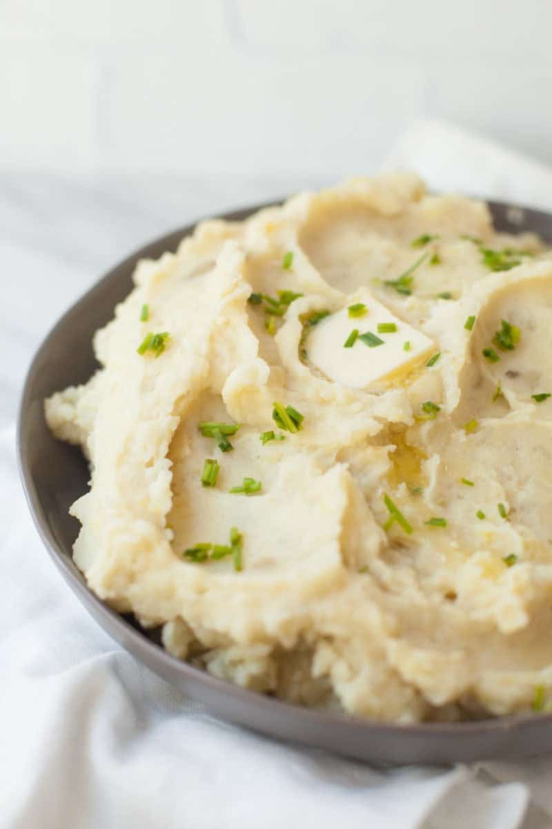 Tight view of bowl of fluffy slow cooker mashed potatoes on white countertop. Mashed potatoes are topped with fresh chives and a melting butter pat.