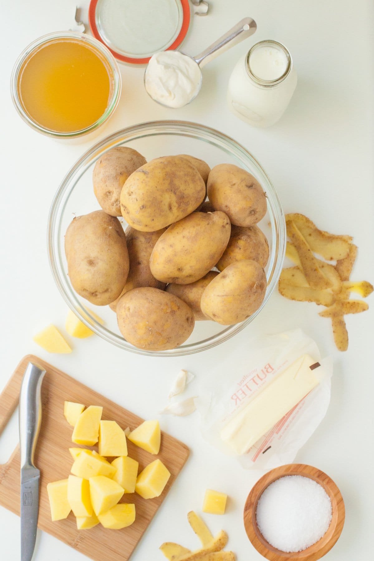 Ingredients (butter, broth, russet potatoes, salt, milk, sour cream) for slow cooker mashed potatoes on white counter top.