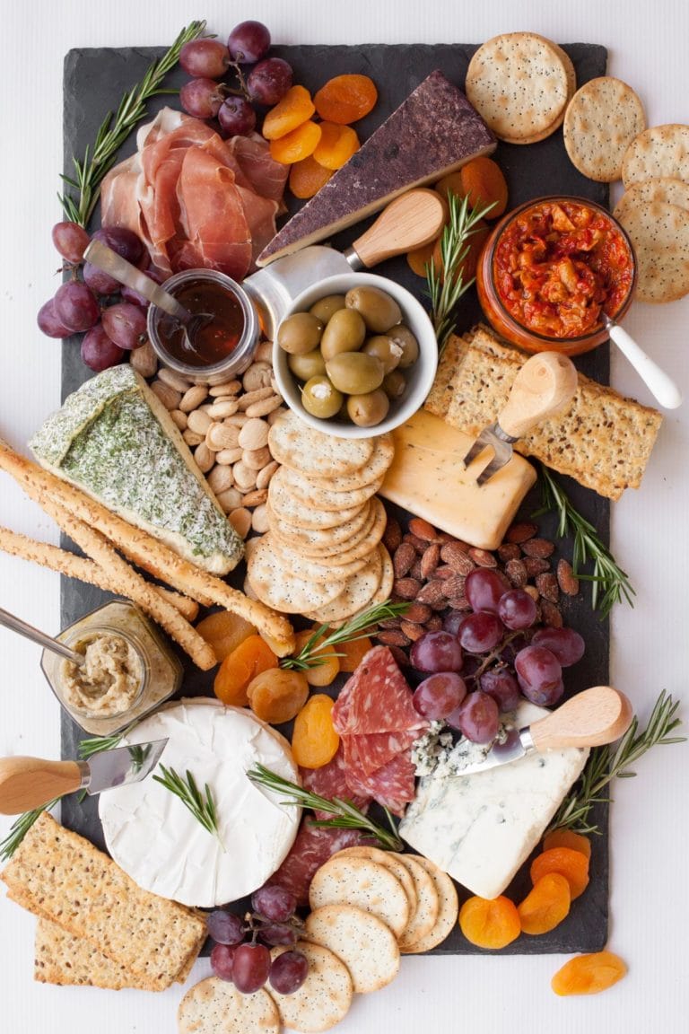 Slate cutting board with 5 wedges of cheese, each with a cheese knife. Various crackers, nuts, dried apricots, olives, condiments, and charcuterie are around the cheese. The board is garnished with fresh rosemary.